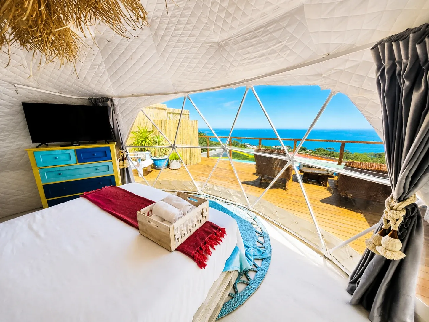 Bed inside a glamping dome facing a panoramic window, a deck and the sea